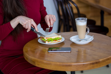 A girl at the table cuts a sandwich at a business lunch, close-up.  Girl with mobile phone and a latte in cafe.の写真素材