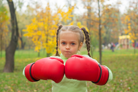 A little girl in huge boxing gloves makes an evil face. A little girl in huge boxing in a park.の写真素材