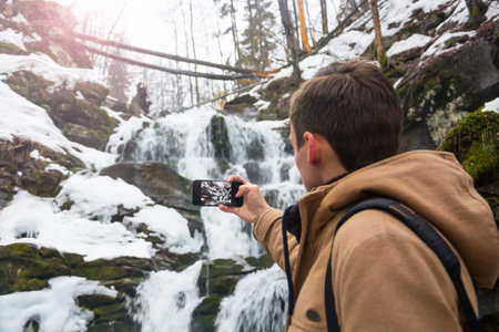 A young man takes a picture of a waterfall on a mobile phone in the spring in the mountains