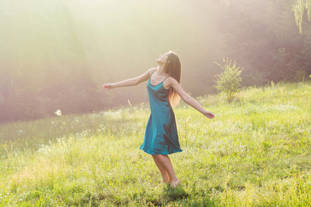 Beautiful smiling young woman is dancing and whirls barefoot on a flower meadow at sunset. Young beautiful woman enjoys sunlight, raising her hands up on a flower meadow near the forest at sunsetの写真素材