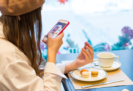 A woman with a smartphone is taking a picture of a macaroons and a cup of tea in a cafe. Food photo.の写真素材