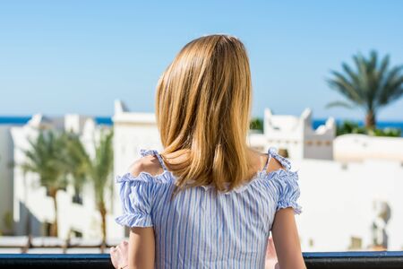 Young girl in sunglasses stands a back on the balcony and looks at the sea and palm trees.の写真素材