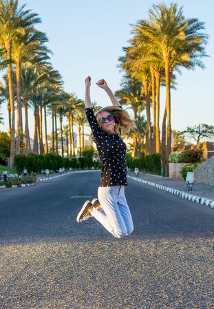Young happy woman in sunglasses is jumping in front of the blooming hibiscus bushes and palm trees at sunsetの写真素材