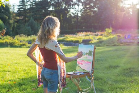 A young woman artist holds a brush and paints a picture on an easel iof the sunset. The painter paints oil paintings in the garden at sunsetの写真素材