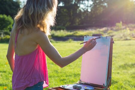 A young woman artist holds a brush and paints a picture on an easel iof the sunset. The painter paints oil paintings in the garden at sunsetの写真素材