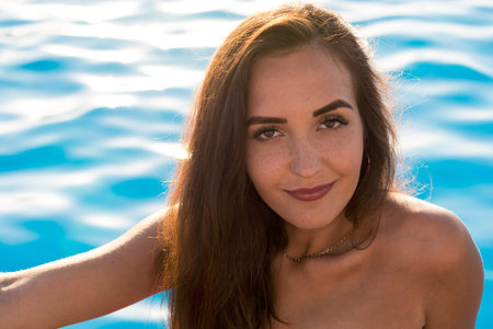 Portrait of a young woman with freckles with long hair in pink bikini sits on the edge near the pool...