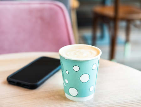 Mint green disposable paper cup of coffee and black cellphone on light wooden table in shopping mall cafe. Lifestyle concept.の写真素材