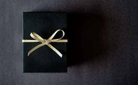 Black gift box with light golden metallic ribbon on dark background. Flat lay minimal style.の写真素材