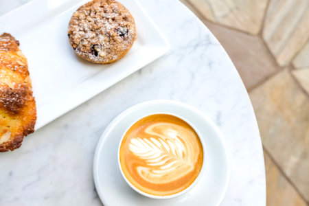 White cup of coffee latte and plate with freshly baked croissant and blueberry tart on marble table...