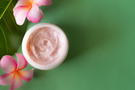 Pink strawberry scrub or cream and Hawaiian plumeria tropical flowers on dark green background. Skin...