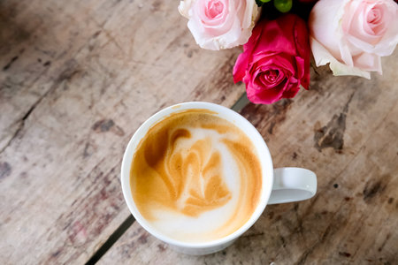 White cup of coffee latte and pink roses on wooden table. Lifestyle concept.