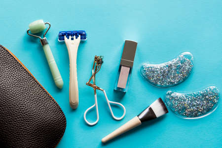 Brown cosmetic bag with lip gloss, jade roller, brush and eyelash curler on turquoise background. Beauty and makeup concept. Flat lay design.の写真素材