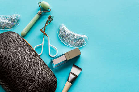 Brown cosmetic bag with lip gloss, jade roller, brush and eyelash curler on turquoise background. Beauty and makeup concept. Flat lay design.の写真素材