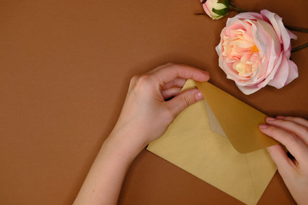Golden envelope, artificial pink rose and woman's hand on brown background with golden confetti...