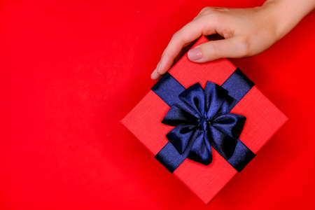 Red gift box with dark blue shiny bow in woman' hand on vivid background. Festive flat lay concept.の写真素材