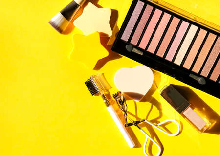 Assorted cosmetic products, eye shadows palette, lip gloss, blow comb, eyelash curler, brush and sponges on yellow background. Beauty concept. Flat lay design.の写真素材