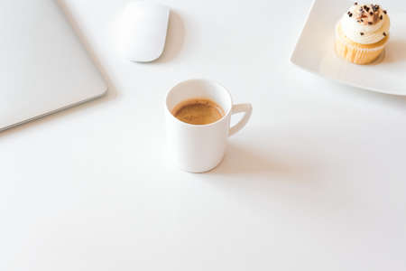 Cup of coffee and cupcake, silver laptop on white desk. Coffee break in home office concept. の写真素材