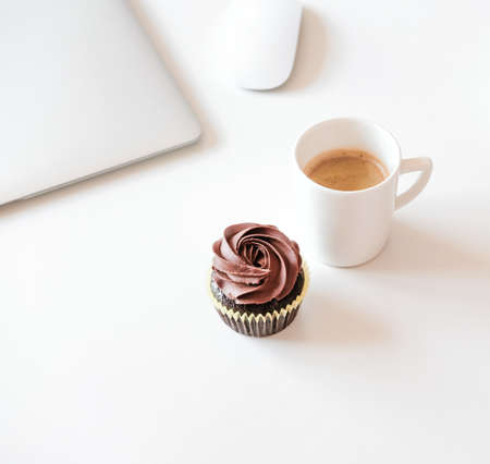 Cup of coffee and chocolate cupcake, silver laptop on white desk. Coffee break in home office concept. の写真素材