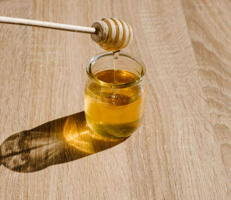 Honey in glass jar and wooden honey dipper on light wooden table background. の写真素材