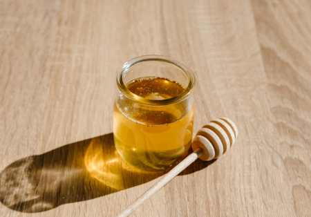 Honey in glass jar and wooden honey dipper on light wooden table background. 