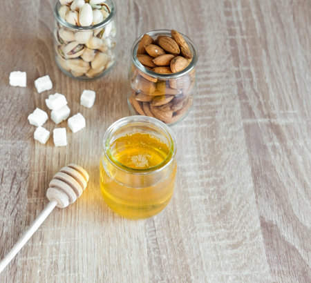 Honey in glass jar with wooden honey dipper and almonds, peanuts on light wooden table background. 