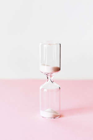 Hourglass on pink and white background, sand flowing through the bulb of sandglass measuring time. minimal concept.の写真素材