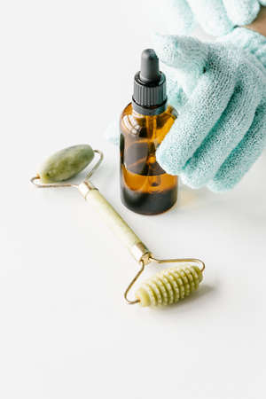 Brown plastic bottle for liquid cosmetics in womans hands in green gloves, green jade massage roller on white table. Cosmetologist holding spin care products.の写真素材