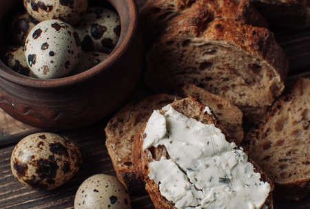 dark buckwheat bread is spread with cottage cheese with herbs in a cut on a wooden table near quail eggs in a clay plate in a rustic style. Snack and breakfast concept.の写真素材