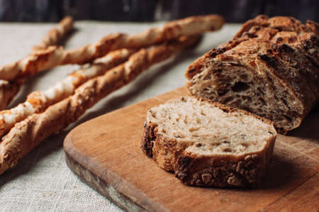 dark yeast-free buckwheat bread in a cut lies on a cutting wooden board on a wooden table, next to it is Italian grissini on a linen tablecloth in a rustic style. Breakfast cooking concept.の写真素材