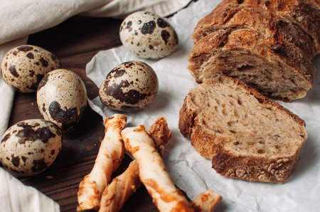 set dark yeast-free buckwheat bread in a cut lies on parchment, next to quail eggs and Italian grissini on a linen tablecloth on a wooden table in a rustic style. Breakfast cooking concept.の写真素材
