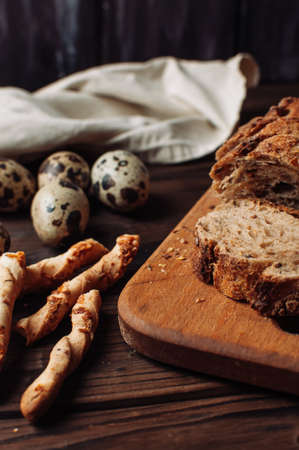 set dark yeast free buckwheat bread in a cut lies on a chopping wooden board, next to quail eggs and Italian grissini on a linen tablecloth on a wooden table in rustic style.Breakfast cooking concept.の写真素材