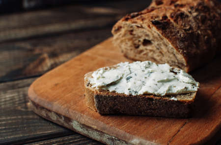 dark bread is spread with cottage cheese with herbs in a cut on a wooden board on a wooden table in a rustic style. Snack and breakfast concept. Selective focus.の写真素材