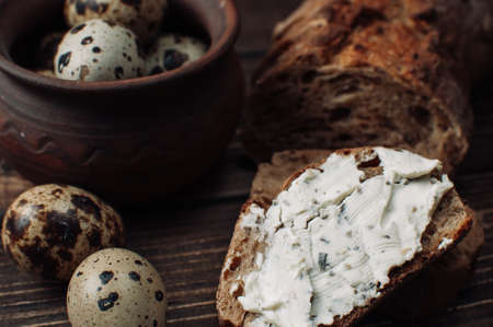 dark buckwheat bread is spread with cottage cheese with herbs in a cut on a wooden table near quail eggs in a clay plate in a rustic style. Snack and breakfast concept.の写真素材