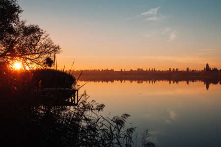 Orange sunset against the background of the river in September in the harvest season. Reflection in the water from the trees. Beautiful autumn landscape. Autumn colors are reflected in calm waters.の写真素材