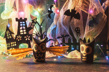 Witch's house with a grave and a scary tree cut out of black paper, next to it there are owls on a wooden wall with a cobweb and LED garland. Copy space. Crafts for halloween celebration concept.の写真素材