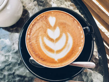 Top view on hot latte coffee with latte art in a vintage frosted black cup and saucer on a glass table with a foam pattern. Image stacking methods.の写真素材