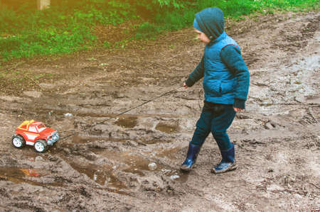 a child in a blue yolk and rubber boots carries a typewriter by a rope through the mud. A child walks outside after a summer rain. A concept of a carefree childhood.の写真素材