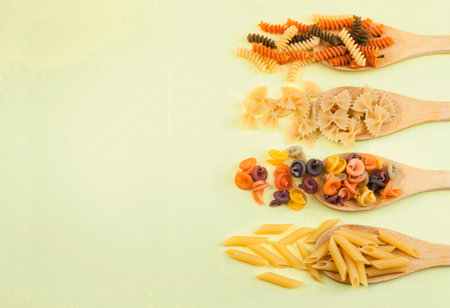 various pasta of Italian cuisine. Farfalle, penne rigate, fusilli on wooden spoons on a yellow...
