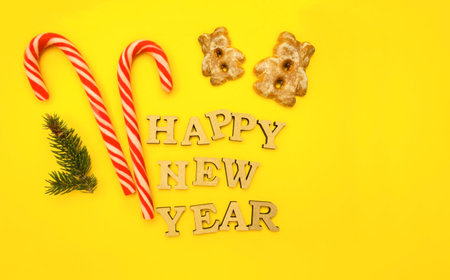 Celebrating Happy New Year. Wooden letters in a New Year's cap on a yellow background with red sweets, gingerbread. Festive decoration or postcard concept. Top view and flat. Copy spaceの写真素材