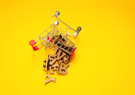 scattered wooden letters of the English alphabet from a miniature shopping trolley on a yellow...