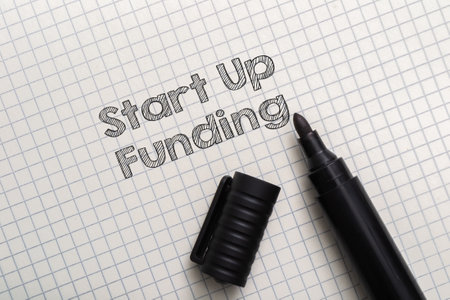 Composition with text Start Up Funding and black marker on checkered paper sheet background upper view. Startup capital for launching new businessの写真素材