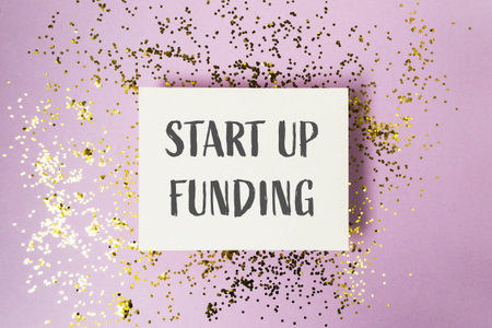 A white sign with the words start up funding written on it. The sign is surrounded by glitter, giving it a festive and celebratory feelの写真素材