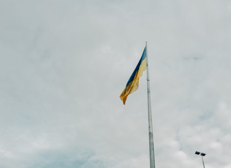 A flag with a yellow stripe and blue stripe is flying high in the sky. The sky is cloudy and the sun...