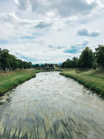 A river with a bridge in the background. The water is murky and the sky is cloudy. Scene is somewhat...
