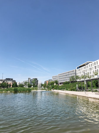 A calm body of water with a clear blue sky above. The water is surrounded by a park and buildings
