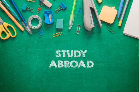 A green chalkboard with a bunch of office supplies and the word study abroad written on it. The supplies include scissors, pens, and a staplerの写真素材
