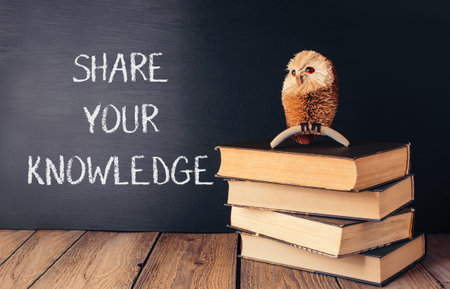 A chalkboard with the words share your knowledge written on it. A small owl figurine is sitting on top of a stack of booksの写真素材