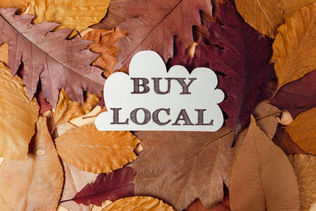 A white sign that says Buy Local is on top of a pile of leaves. Concept of autumn and the idea of supporting local businessesの写真素材