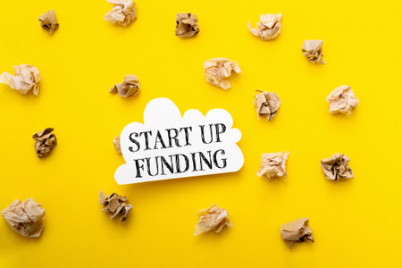 A yellow background with a white sign that says Start Up Funding. The sign is surrounded by shredded paperの写真素材