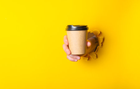 Person is holding a coffee cup with a lid in their hand, and the cup is partially cut out of a yellow background. Concept of surprise or curiosityの写真素材